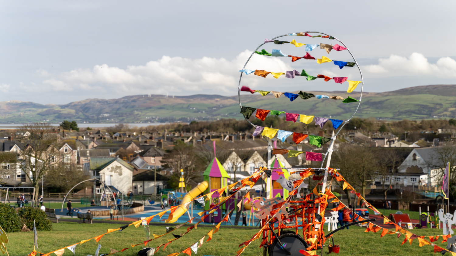 A small cart with multi-coloured flags and bunting hanging off it sat on top of Millom Park. Behind the park you can see houses and the hills.