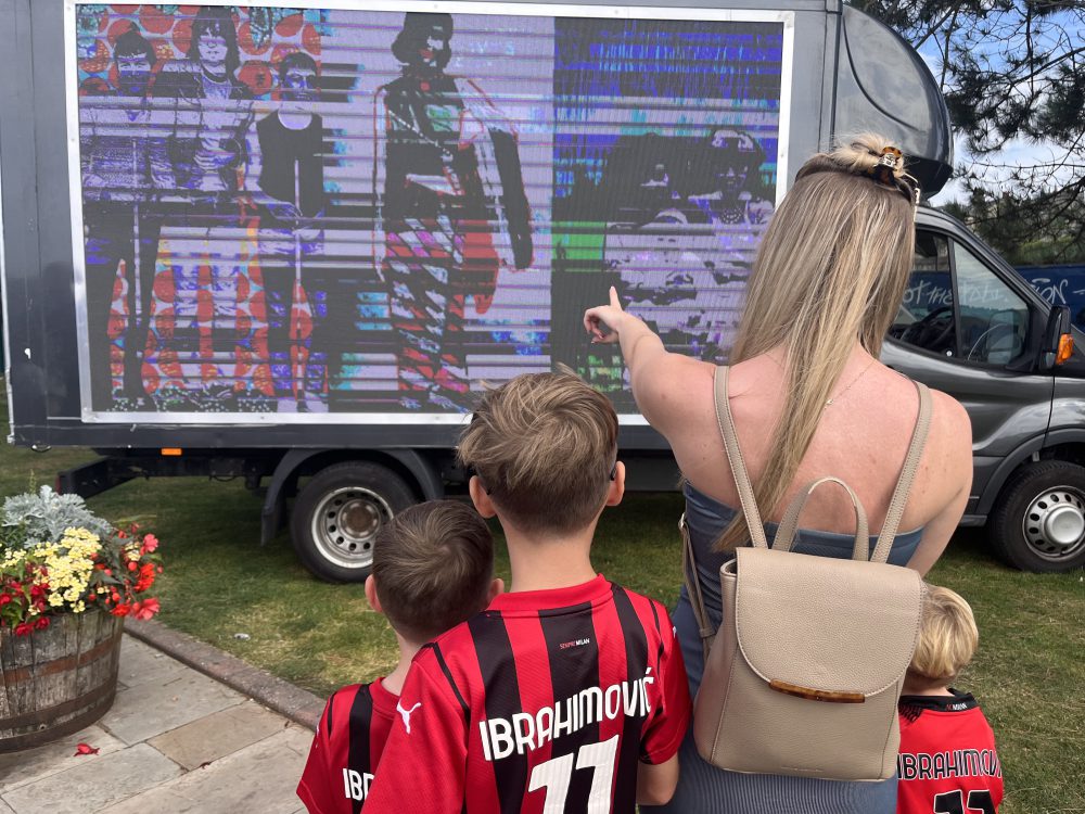 A woman and group of children gather infront a van with a big screen on it. The woman is pointing at the screen.