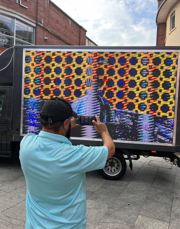 A woman and group of children gather infront a van with a big screen on it. The woman is pointing at the screen.